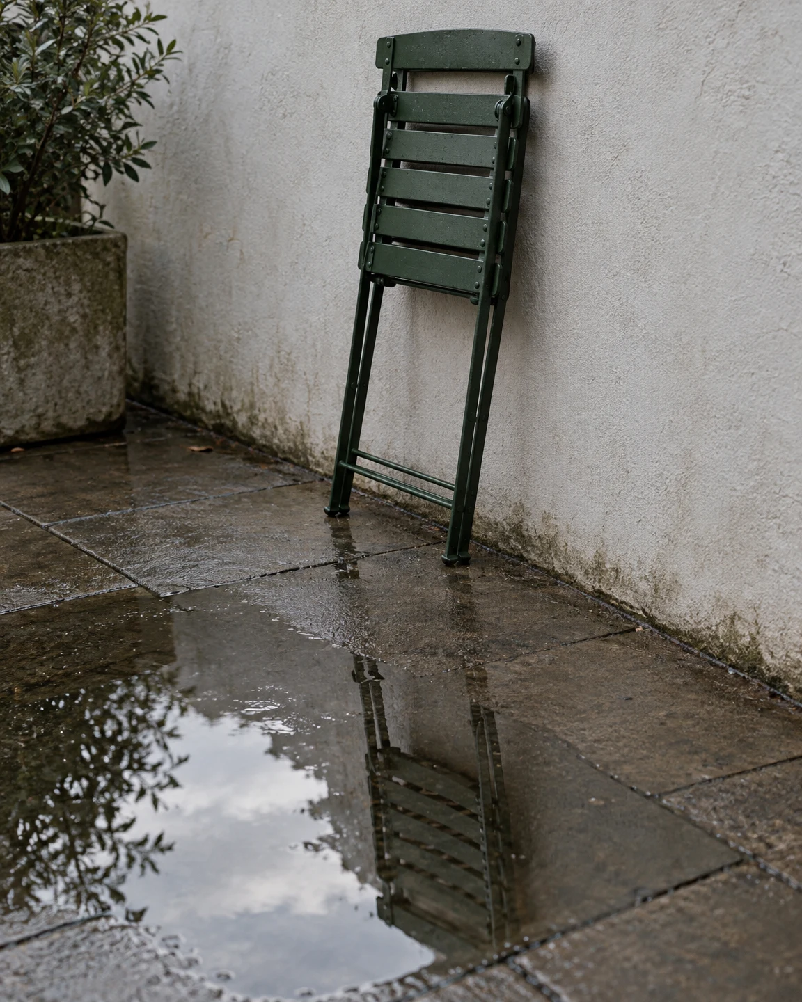A folded dark-green metal patio chair leans against a pale textured wall above wet stone paving, with its reflection visible in a puddle; a leafy plant in a…