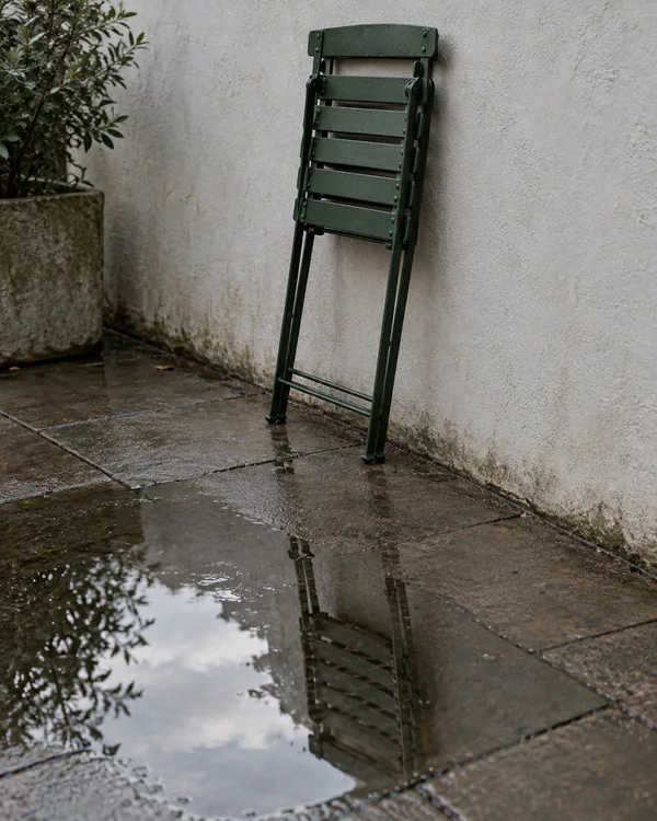 A folded dark-green metal patio chair leans against a pale textured wall above wet stone paving, with its reflection visible in a puddle; a leafy plant in a…