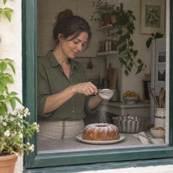 A woman in a muted green shirt sifts powdered sugar over a golden ring cake on a plate, seen through a dark window screen in a leafy kitchen.