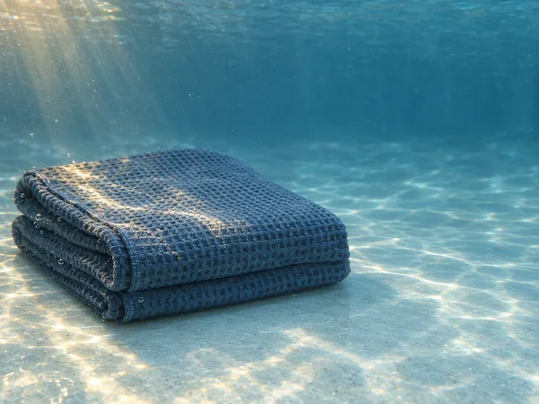 A folded slate-blue waffle-weave towel rests on the floor of a blue underwater tank, with warm light crossing its top and front edge and open water to the…