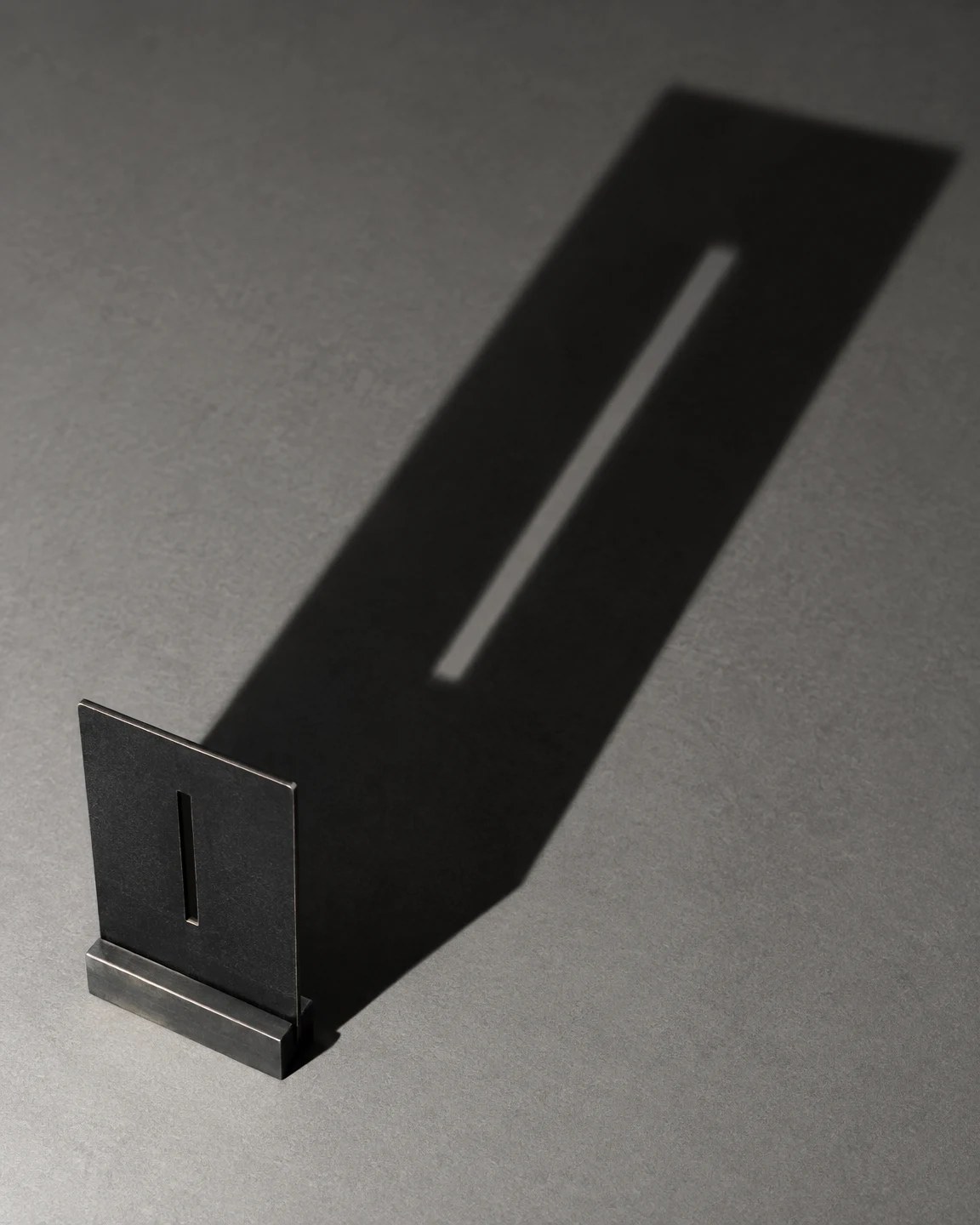 A matte-black rectangular steel plate with a narrow vertical slot stands in a metal foot on a gray surface, casting a long diagonal shadow with a bright strip…