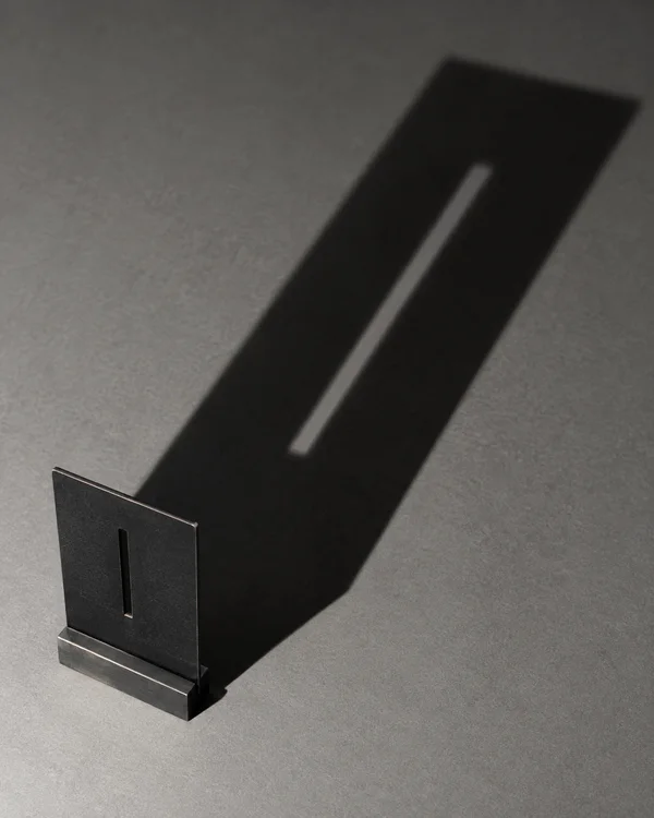 A matte-black rectangular steel plate with a narrow vertical slot stands in a metal foot on a gray surface, casting a long diagonal shadow with a bright strip…