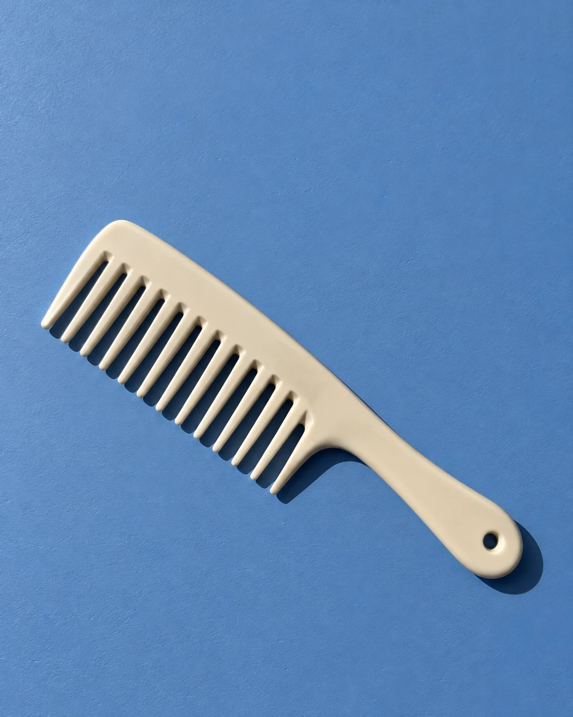 An ivory wide-tooth comb lies diagonally across a textured blue tabletop, with a crisp dark shadow extending to the right and open space above it.