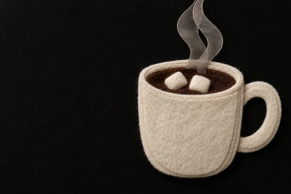 A cream wool-felt mug filled with dark cocoa and two marshmallows sits against a textured black fabric background, with two translucent gray steam ribbons…