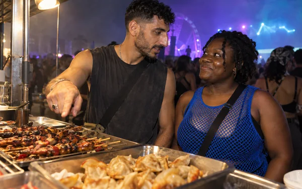 A man points toward trays of grilled skewers and dumplings at a food counter while a woman in a blue mesh top smiles at him; blurred festivalgoers and violet…