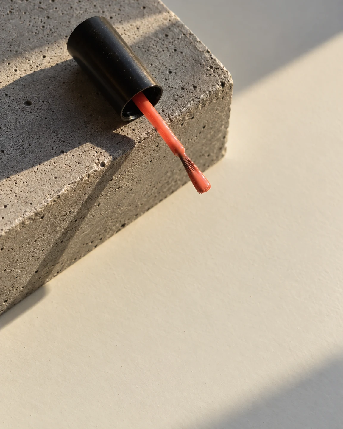 A black cylindrical nail-polish cap rests on a grey concrete block, with a coral applicator extending diagonally over the off-white tabletop and its bristle…