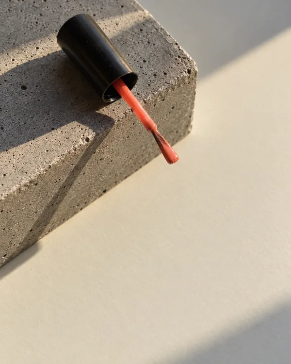 A black cylindrical nail-polish cap rests on a grey concrete block, with a coral applicator extending diagonally over the off-white tabletop and its bristle…
