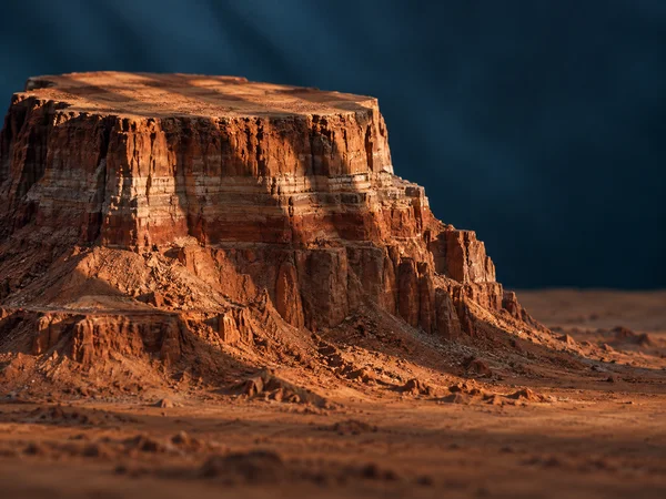 A broad, flat-topped rust-orange mesa rises from a rocky foreground, lit warmly against a softly blurred deep blue background with open space on the right.