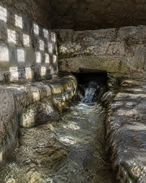 Clear water spills from a dark opening in a weathered limestone springhouse and runs through a shallow stone channel toward the foreground.