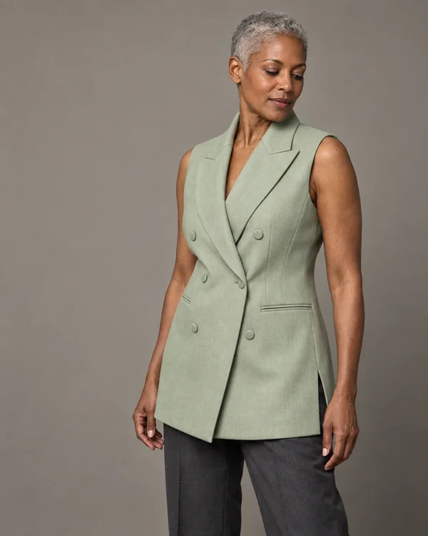 A woman with closely cropped silver hair wears a pale green double-breasted longline waistcoat with peaked lapels and welt pockets over dark trousers, standing…