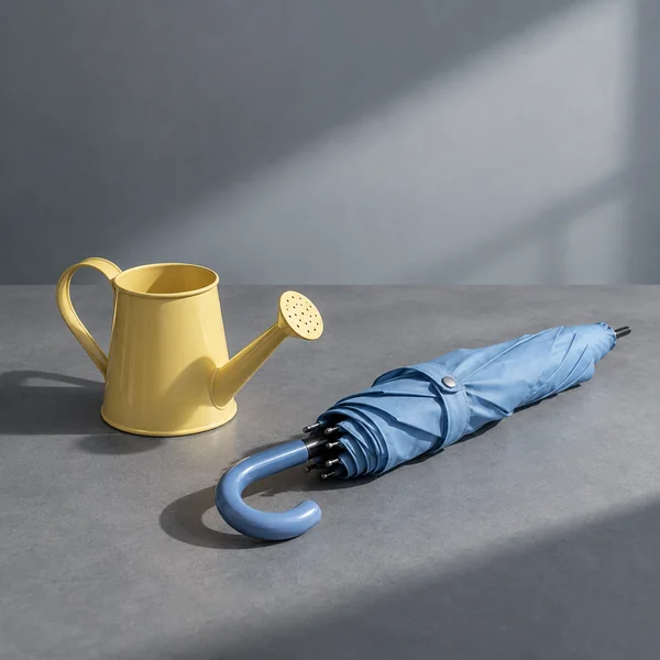 A butter-yellow metal watering can and a folded sky-blue umbrella with a curved handle sit on a grey tabletop beneath a broad shaft of light, with generous…