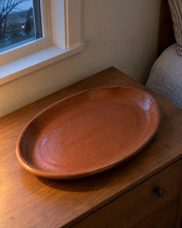 An empty oval terracotta platter rests diagonally on a wooden chest beneath a window in a bedroom corner, with cool dusk light at the window and warm light…