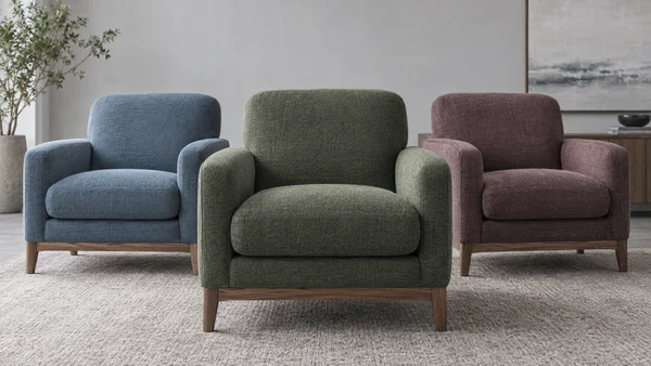 Three upholstered armchairs in evergreen, dusty blue, and muted mauve sit on a pale rug in a gray-walled room; the green chair is centered slightly forward…