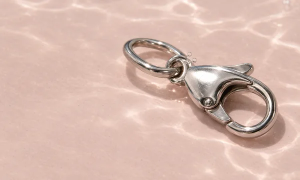 A polished silver lobster clasp with an oval jump ring lies diagonally on a pale blush surface under clear water, with small bubbles near its hinge.
