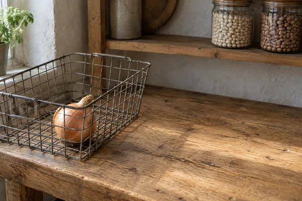 A brown onion sits in the front corner of a grey wire basket on a wooden pantry workbench, with jars of dry beans on a shelf behind and a potted plant beside…