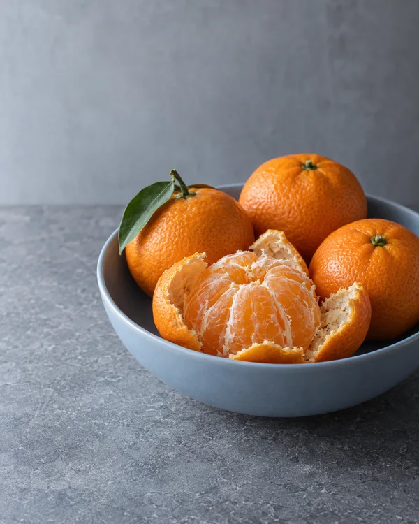 Three whole mandarins and a partly peeled mandarin sit in a pale blue ceramic bowl on a grey stone surface, against a softly textured grey background.