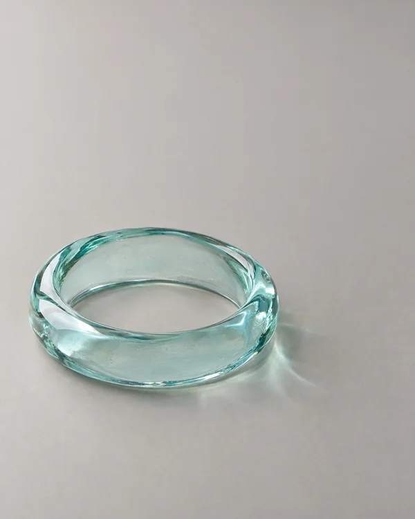 A thick pale-aqua glass bangle rests on a matte warm-grey surface, with its oval opening visible and a faint tinted light pattern beside it.