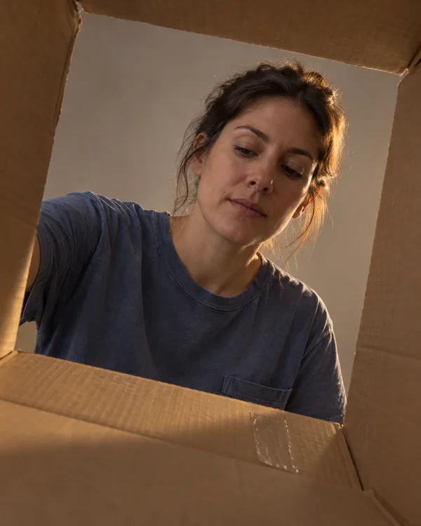 A woman in a faded blue shirt leans over an open cardboard moving box, looking down inside it against a warm-grey studio backdrop; amber light rims her hair…