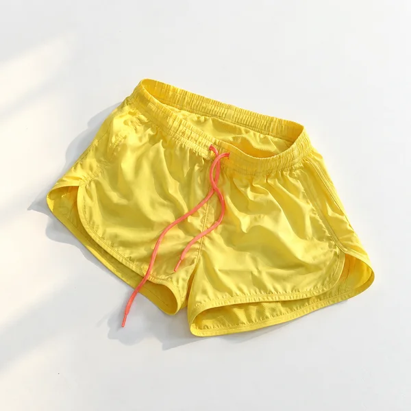 Overhead view of lemon-yellow running shorts with a coral drawstring, casually spread across a matte-white surface.