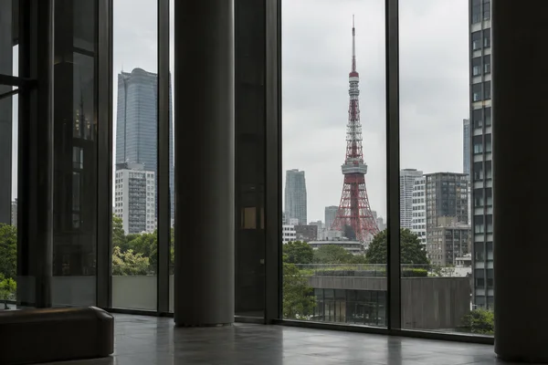 Tokyo Tower rises above trees and surrounding high-rises, viewed through the tall windows of a shadowed atrium with dark columns and a stone floor in the…