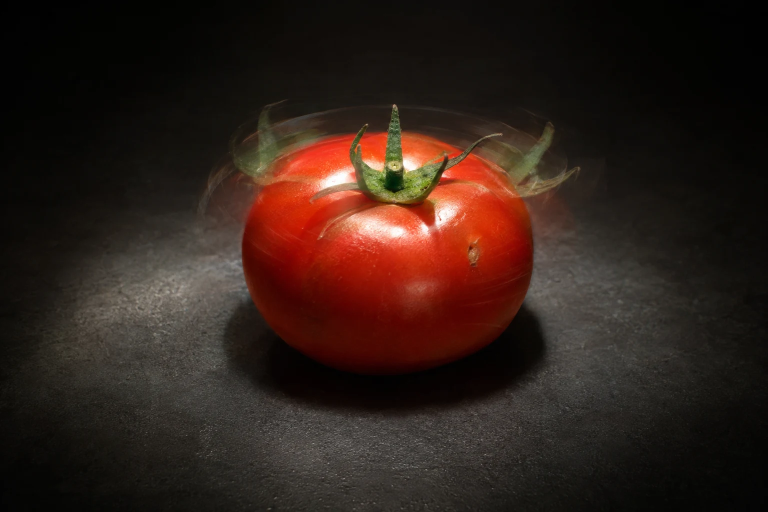 A ripe red tomato sits on a charcoal tabletop under a focused pool of light, with a crisp green calyx and faint translucent calyx positions around it.
