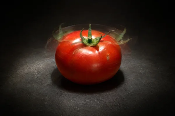 A ripe red tomato sits on a charcoal tabletop under a focused pool of light, with a crisp green calyx and faint translucent calyx positions around it.
