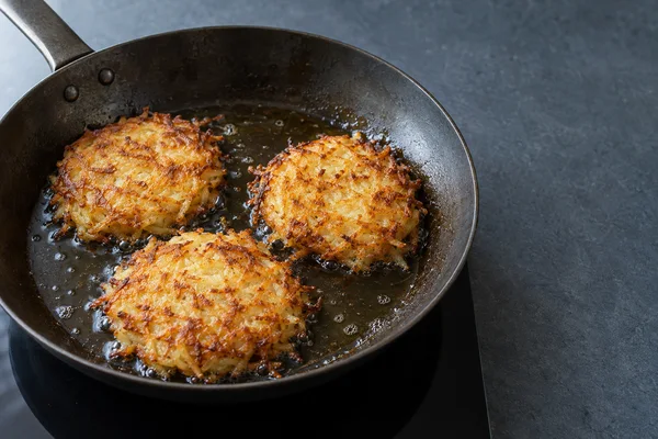 Three golden shredded-potato rösti fry in a dark skillet on a black induction hob, with bubbling oil around them and clear blue-grey counter space to the right.