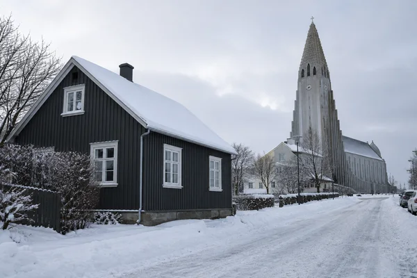 A snow-covered residential street in Reykjavík with a dark timber cottage in the left foreground and Hallgrímskirkja rising behind nearby houses and bare trees…