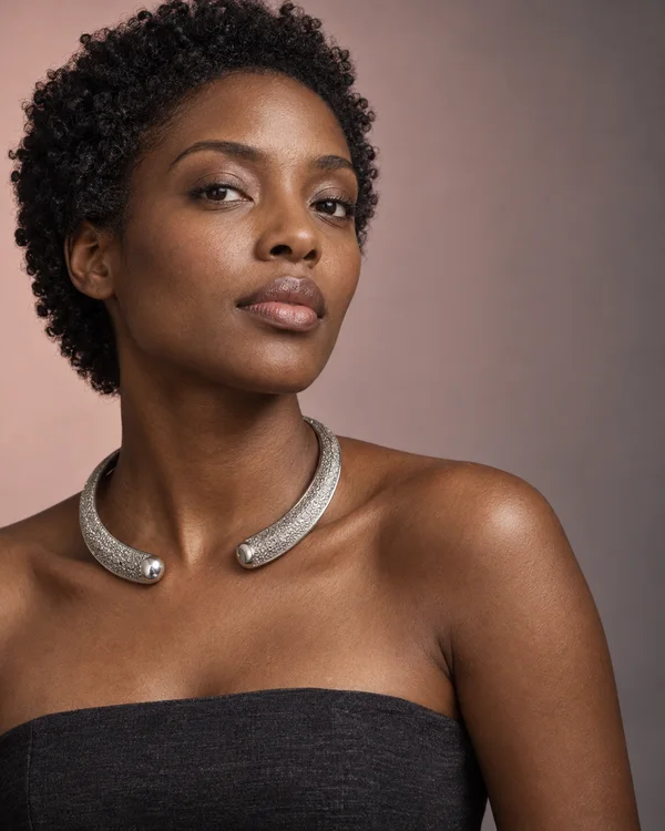 A woman with deep brown skin and short natural curls wears a textured silver open-ended collar and a charcoal strapless top against a muted pink-to-grey studio…