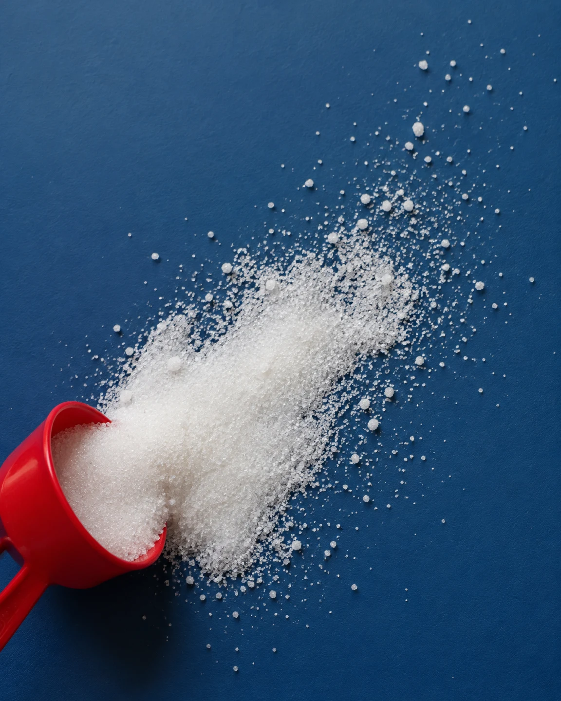 A red plastic measuring scoop lies tipped at the lower-left of a deep-blue surface, spilling a broad diagonal trail of white sugar toward the upper-right.