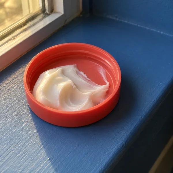 A plain coral-red plastic jar lid rests inner-side up on a cobalt-blue painted windowsill, holding a glossy curl of white cream.