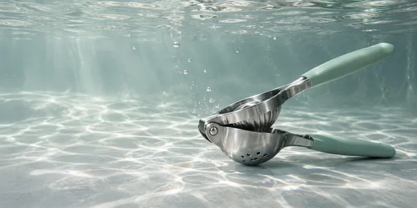 A stainless-steel citrus press with muted seafoam grips rests open at an angle on a pale tank floor underwater, with light caustics and bubbles around it.