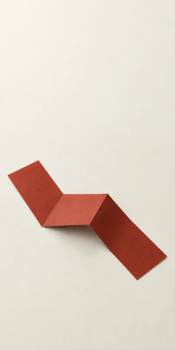 A rust-red paper strip folded into a three-segment zigzag lies diagonally across a warm ivory textured background, with a soft shadow beneath the raised middle…