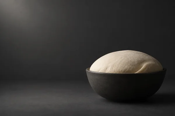A pale ivory dome of dough with a folded edge rises from a low charcoal bowl on a dark, subtly textured surface, with open space to the left.