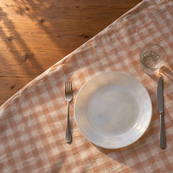 Overhead view of a white pearlescent dinner plate with a fork, knife and clear tumbler on a faded peach-and-cream gingham tablecloth, with warm wooden boards…