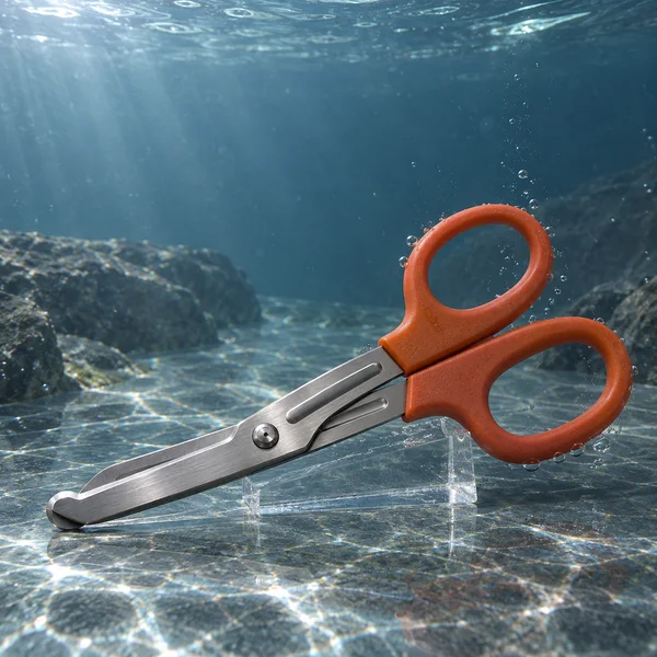 Large orange-handled stainless-steel shears rest diagonally on a transparent wedge above a textured rocky floor underwater, with bubbles around the handles and…
