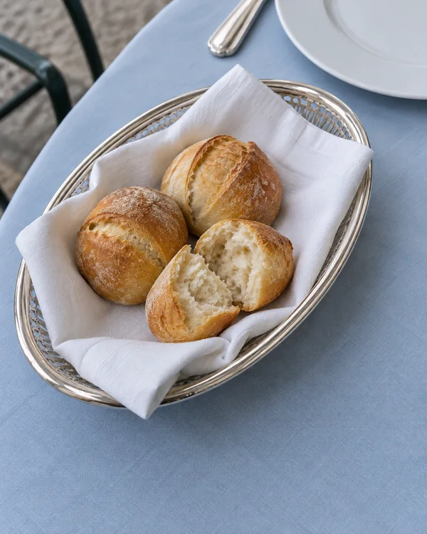 An oval silver bread basket lined with white linen holds two intact golden rolls and the separated halves of a third, showing its pale interior.
