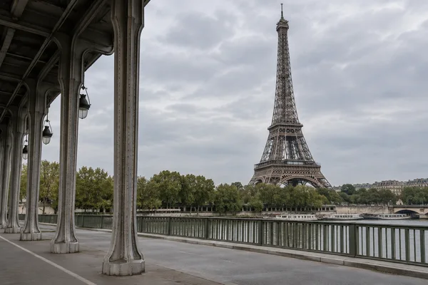 The Eiffel Tower rises beyond the tree-lined Seine and opposite embankment, framed by iron columns and the underside of a viaduct along a quiet pedestrian…