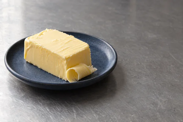 A pale yellow rectangular block of butter with fine ridges and a curled shaving sits on a dark blue ceramic plate on a scratched grey metal tabletop, with open…