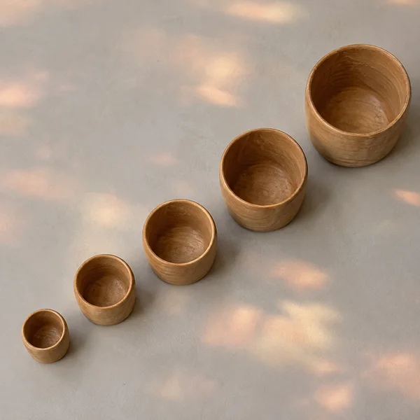 Five unpainted honey-brown wooden cups of increasing size are arranged diagonally on a pale gray surface, with soft amber and pinkish patches of light across…