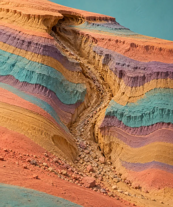 A close view of a deep runoff channel cutting through a layered, brightly banded eroded riverbank, with loose stones collected along its base and a turquoise…