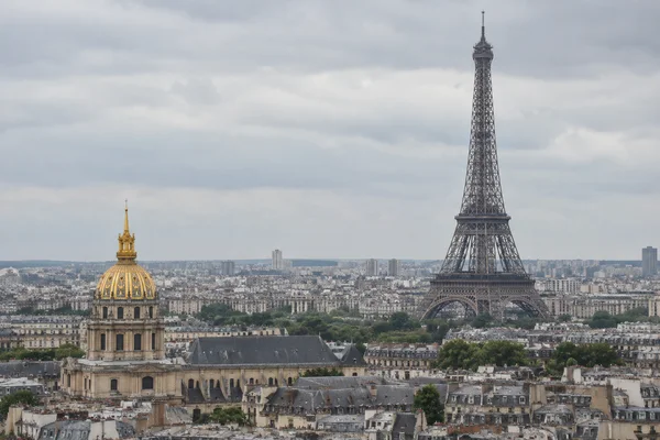 An elevated view across Paris rooftops shows the Eiffel Tower rising on the right and the gilded dome of Les Invalides in the left foreground beneath a pale…