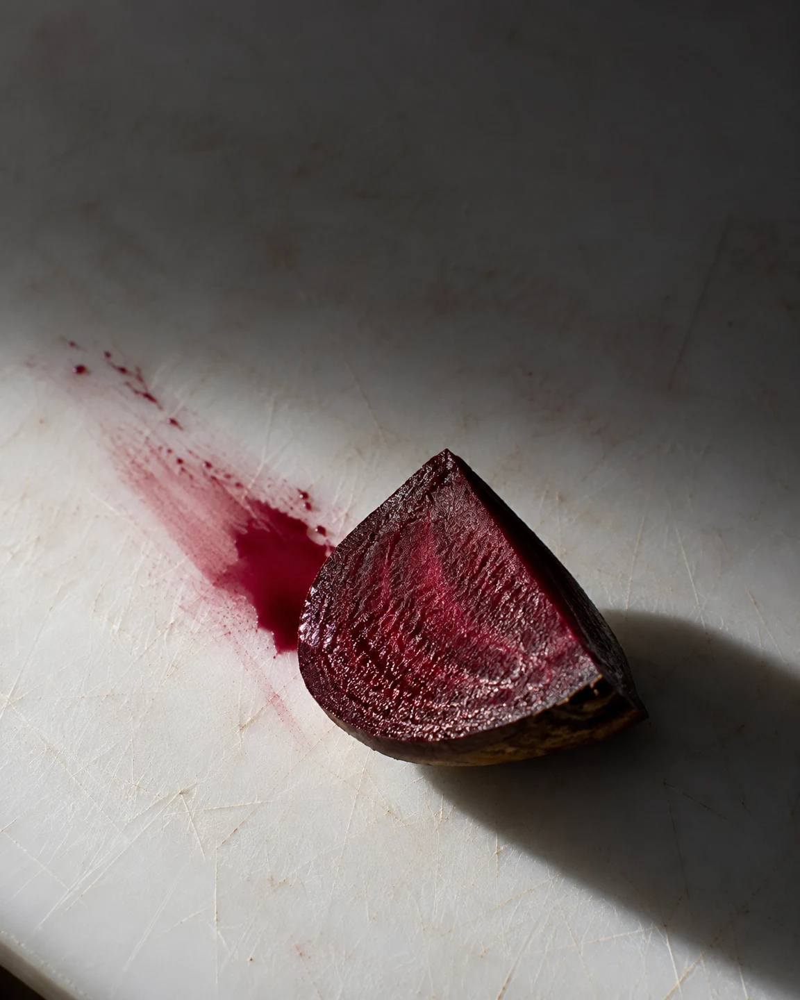 A freshly cut beet wedge rests on a heavily scratched white cutting board, its glossy burgundy face beside a diagonal crimson smear and pooled juice.