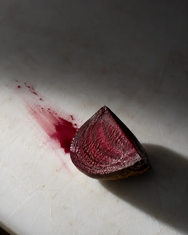 A freshly cut beet wedge rests on a heavily scratched white cutting board, its glossy burgundy face beside a diagonal crimson smear and pooled juice.