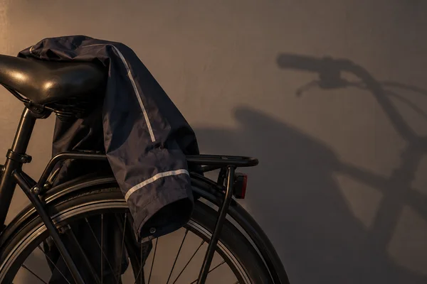 A navy raincoat with silver reflective trim draped over the saddle and rear rack of a black bicycle, beside its cropped rear wheel.