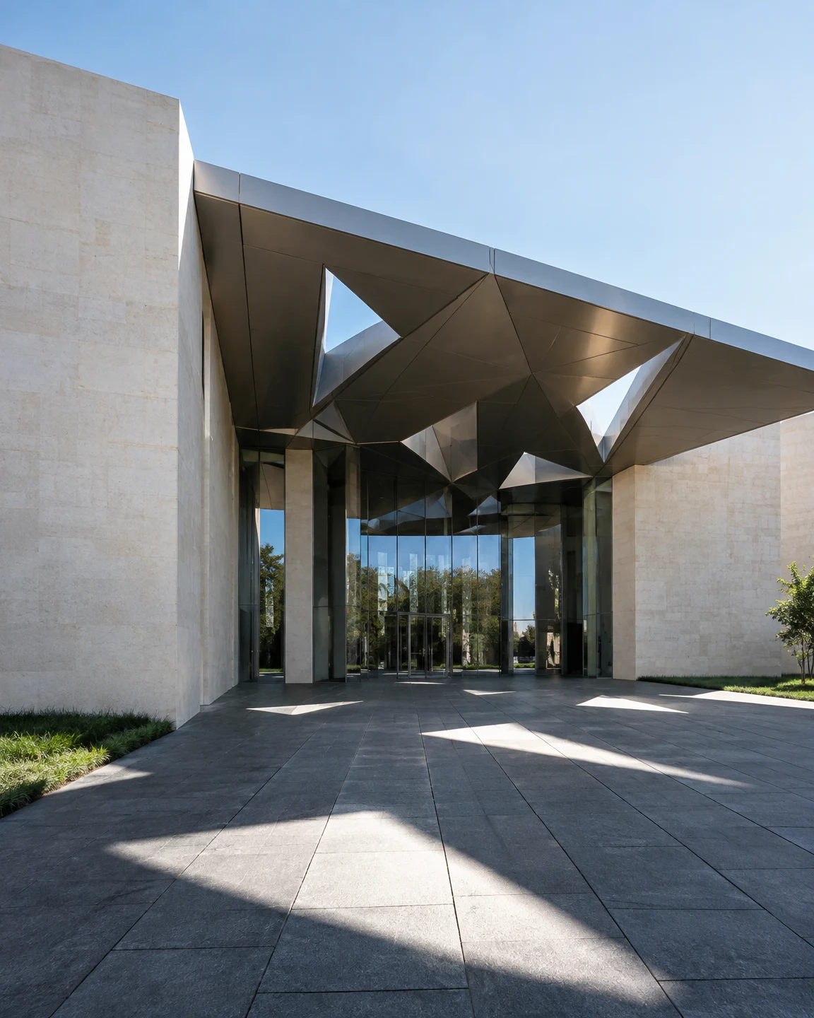 A contemporary pale-stone building with a tall glass entrance and angular metal canopy, seen from a broad charcoal-paved forecourt under a clear blue sky.