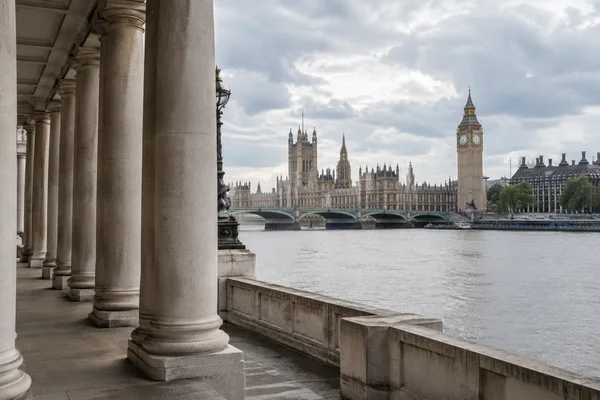 A row of pale stone columns and a riverside parapet frame the Thames, with the Palace of Westminster and Elizabeth Tower visible across the water beneath a…