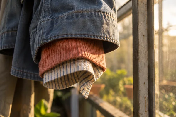 Close-up of a faded blue work-jacket sleeve layered over a coral ribbed sweater and cream-and-blue striped shirt, hanging beside a sunlit greenhouse window.