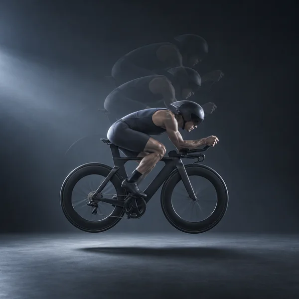 A helmeted cyclist in a dark sleeveless suit rides a black time-trial bicycle in a low aerodynamic tuck, suspended above a charcoal studio floor with a…
