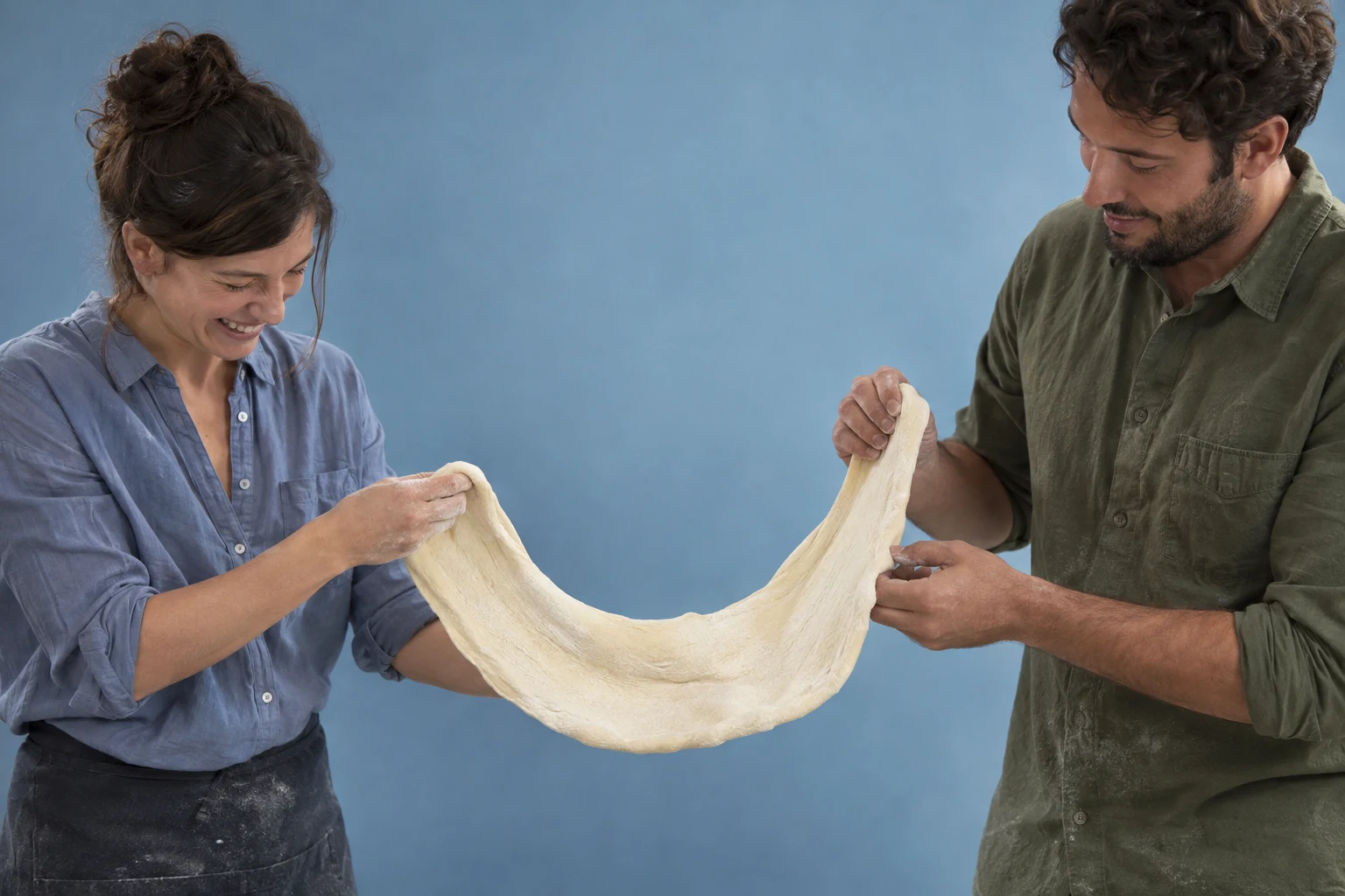 Two adults in casual shirts stretch a broad sheet of pale dough between them against a dusty-blue studio backdrop.